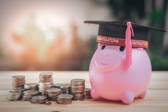 piggy bank with a graduation cap beside a stack of coins for tuition